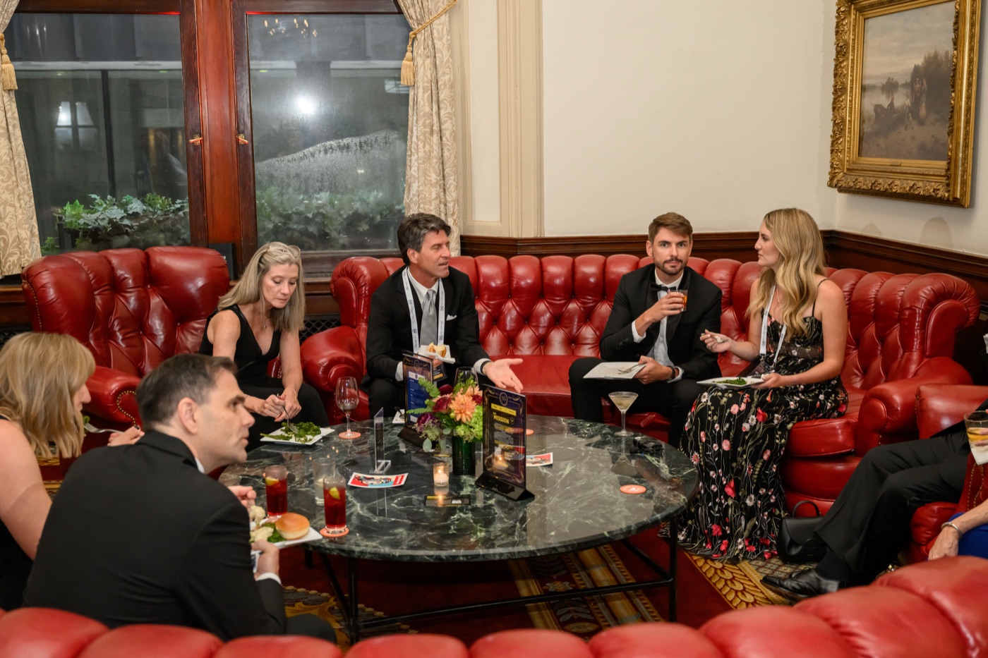 Five guests in black-tie attire in close conversation around a circular table in a chandelier-lit, oak-panelled room.